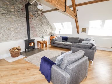 A living room with a stone wall wood stove gray sofas and wooden beams at Yr Hen Festri in Y Felinheli