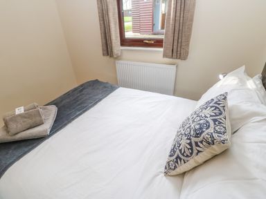 A bedroom with a made bed a patterned pillow folded towels on the bed a window with curtains and a radiator below