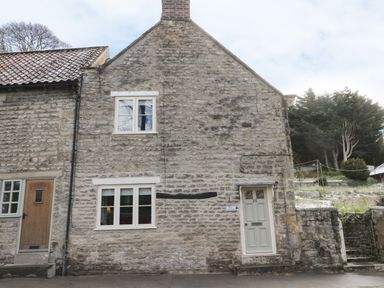A stone cottage with two doors and two windows next to a grassy area with trees at Beck Cottage in Pickering