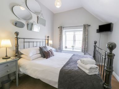 A bedroom with a metal bed frame, bedside tables with lamps, a window with curtains, mirrors on the wall and a wall-mounted television at Beck Cottage in Pickering