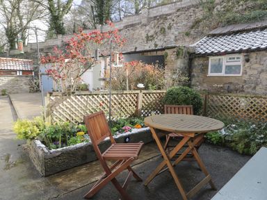 An outdoor patio with a wooden table and two chairs near a raised flower bed and stone wall at Beck Cottage in Pickering