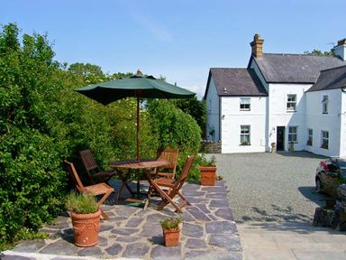 An outdoor patio with wooden table and chairs under a green umbrella near a white house with a gravel driveway at Carreg Rhys in Malltraeth near Newborough