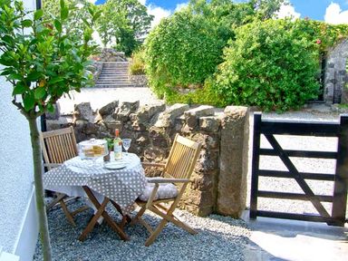 A small outdoor seating area with two wooden chairs and a table with a tablecloth and drinks near a stone wall and gate at Carreg Rhys in Malltraeth near Newborough