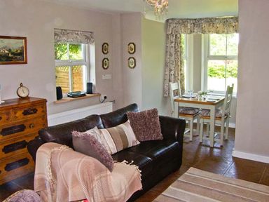 A living room with a dark leather sofa, wooden chest of drawers, and a small dining table near windows at Carreg Rhys in Malltraeth near Newborough