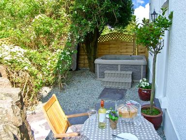 A small outdoor garden area with a hot tub, wooden fence, table with a checked tablecloth, bottle of wine, cake, chairs, and potted plants at Carreg Rhys in Malltraeth near Newborough