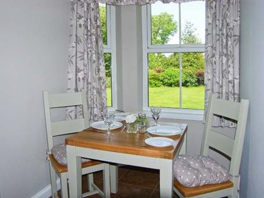 A small dining table with two chairs and place settings by windows with curtains showing a garden at Carreg Rhys in Malltraeth near Newborough