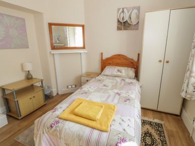 A single bed with floral bedding and folded towels in a bedroom with a wardrobe a bedside table a small cabinet with a lamp and two framed pictures at Bangor Cottage in Bangor