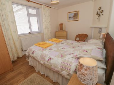 A bedroom with a double bed covered with a patchwork quilt and two yellow towels on top a wooden dresser with a mirror a wicker chair curtains and a window with blinds Bangor Cottage Bangor