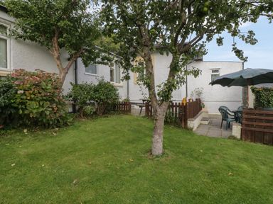 A garden with trees bushes and lawn next to a white house with an outdoor sitting area and umbrella at Bangor Cottage in Bangor