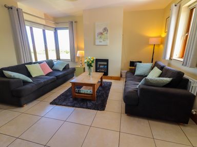A living room with two dark sofas with cushions a wooden coffee table with flowers on a rug and windows with curtains at Brandon Holiday Home in Cloghane County Kerry