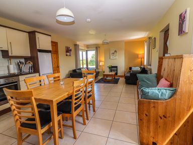 An open plan kitchen and living room with a wooden dining table and chairs two sofas a wooden bench with cushions and lamps at Brandon Holiday Home in Cloghane County Kerry