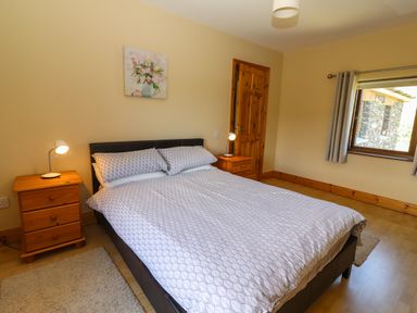 A bedroom with a double bed, two wooden bedside tables with lamps, a window with curtains and a door at Brandon Holiday Home in Cloghane County Kerry
