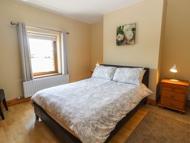 A bedroom with a double bed two bedside tables with lamps a window with curtains and a floral painting on the wall at Brandon Holiday Home in Cloghane County Kerry