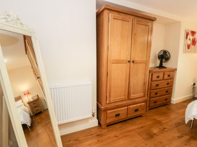 A wooden wardrobe and chest of drawers with a fan on top next to a white radiator and a mirror reflecting a bedside table with a lamp in a bedroom at Ty Taid in Llanddaniel Fab near Beaumaris