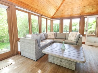 A sunroom with a wicker sectional sofa and a wicker coffee table with a glass bottle on it at Lyons Green Little Fransham near Swaffham