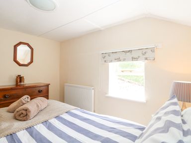 A bedroom with a striped bedspread two rolled towels a wooden dresser with jars a small wall mirror and a window with an animal print blind at Lyons Green Little Fransham near Swaffham