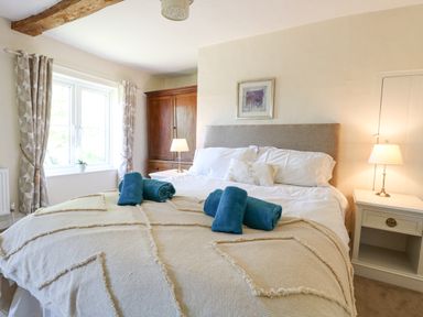 A bedroom with a bed covered with a beige blanket and blue towels a wooden wardrobe by a window with curtains and two bedside tables with lamps at Lyons Green in Little Fransham near Swaffham