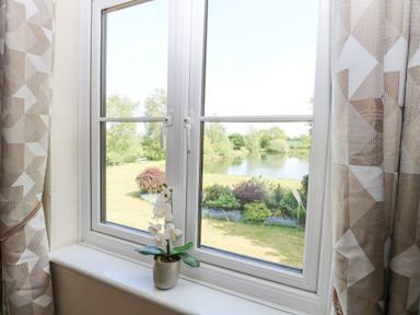 A window with beige geometric patterned curtains and a potted orchid on the windowsill overlooking a garden and a pond at Lyons Green in Little Fransham near Swaffham