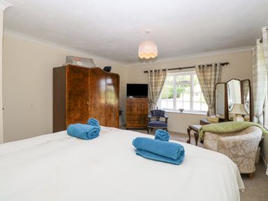 A bedroom with a bed with blue towels on it a wooden wardrobe a TV on a chest of drawers armchairs and windows with curtains at Lyons Green in Little Fransham near Swaffham