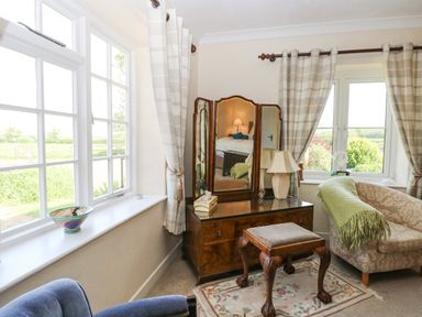 A sitting room with a three-panel mirror on a wooden dresser a cushioned stool a lamp and two windows with curtains at Lyons Green in Little Fransham near Swaffham