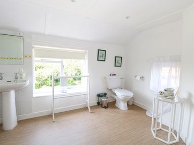 A bathroom with a toilet, a sink, a window with a blind, a small table, and framed pictures on the wall at Lyons Green in Little Fransham near Swaffham
