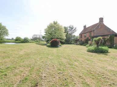A large mowed lawn with trees and shrubs next to a brick house with a chimney near a pond at Lyons Green in Little Fransham near Swaffham