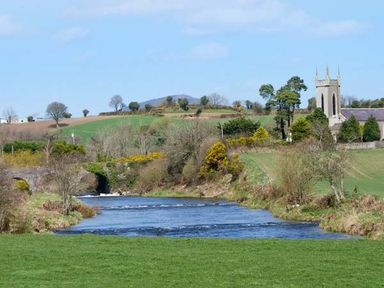 A river flowing through grassy fields with a stone bridge and a church tower in the background at Rosemount Coach House in Ballycarney near Enniscorthy County Wexford