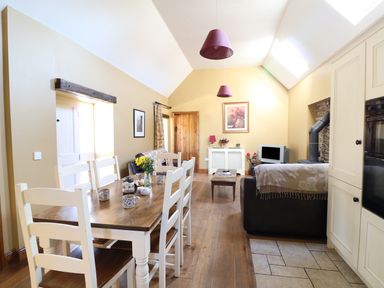 A dining area with a wooden table and six white chairs next to a living area with sofas a television and woodburner at Rosemount Coach House Ballycarney near Enniscorthy County Wexford