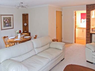 A living room with white sofas a wooden dining table with chairs and a vase with flowers and a kitchen visible through a doorway at 9 Bancroft Place Stratford-upon-Avon