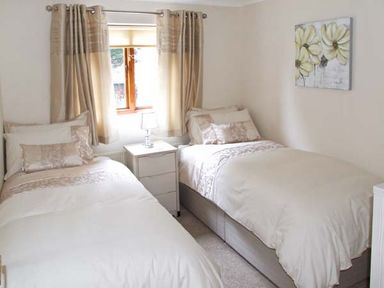 A bedroom with two single beds separated by a small white nightstand with a lamp and a window with beige curtains at 9 Bancroft Place Stratford-upon-Avon