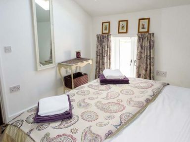 A bedroom with a bed covered with patterned bedding and folded towels a wall mirror a small console table with a basket and framed pictures above a window at Woodstone Barn Whitchurch near Tavistock