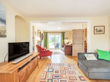 A living room with a gray sofa pillows a patterned rug a wooden cabinet with a television and a red armchair near sliding glass doors at Barafundle House in Tenby
