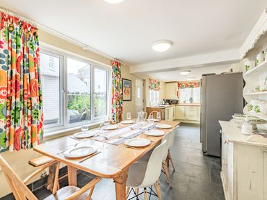 A kitchen with a wooden dining table set with plates and glasses white chairs floral curtains and cupboards at Barafundle House in Tenby