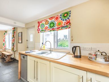 A kitchen with a sink under a window with floral blinds and a wooden countertop with an electric kettle and jars at Barafundle House in Tenby