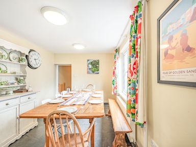 A dining room with a wooden table set for eight with chairs and a bench with colorful curtains and a white cabinet with dishes at Barafundle House in Tenby