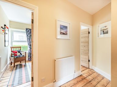 A hallway with wooden floors radiator and framed pictures next to two doorways one leading to a room with a wooden chair and colorful curtains at Barafundle House in Tenby