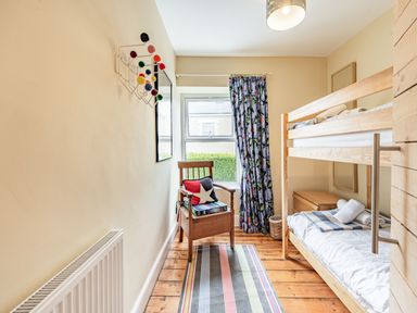 A small bedroom with a wooden bunk bed a striped rug a wooden chair with cushions a window with floral curtains and a colorful wall hook at Barafundle House in Tenby