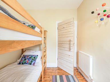 A small bedroom with wooden bunk beds a wooden door a colorful wall hook a radiator and a striped rug on wooden floor at Barafundle House in Tenby