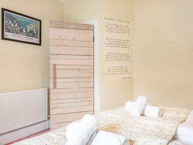 A bedroom with a bed covered in a beige quilt with rolled white towels on it a wooden door slightly open a framed poster on the wall and a poem written on the wall at Barafundle House in Tenby