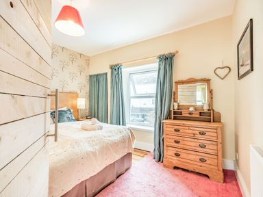 A bedroom with a bed, wooden dresser with mirror, teal curtains, and pink carpet at Barafundle House in Tenby