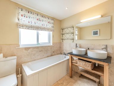 A bathroom with a bathtub toilet two sinks wooden vanity and a window with patterned blind at Barafundle House in Tenby