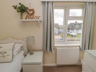 A bedroom with a white bed beside a nightstand with a lamp and a window with curtains at Dune View in Beadnell