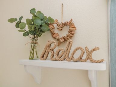 A white shelf with a glass vase holding green leaves and a rope decoration spelling relax hanging wooden heart on wall at Dune View in Beadnell