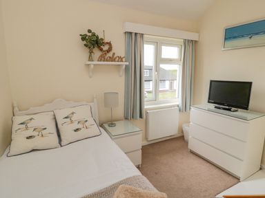 A bedroom with a bed with bird pillows a nightstand with a lamp a window with curtains a dresser with a television and a shelf with decor at Dune View in Beadnell