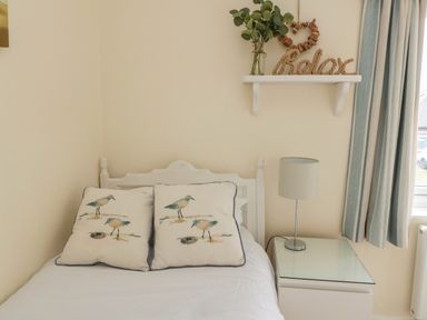 A bedroom with a white bed with bird printed pillows a bedside table with a lamp a white shelf with plants and the word relax and striped curtains at Dune View in Beadnell