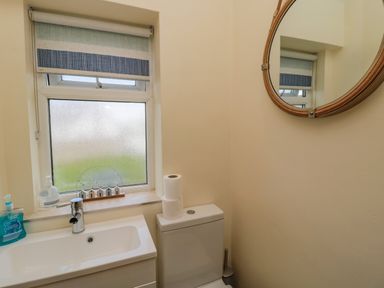 A bathroom with a sink, faucet, hand soap, toilet with toilet paper rolls, window with a striped blind, and a round wall mirror at Dune View in Beadnell