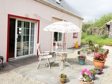 An outdoor patio with a round table and two chairs under an umbrella and potted plants near a house with sliding glass doors at Fuschia Cottage in Waterville County Kerry