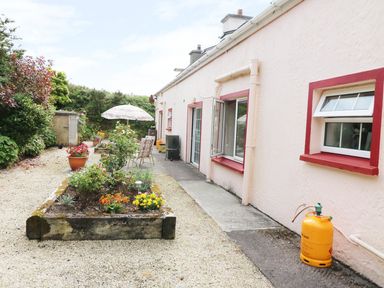 An outdoor garden area with chairs and umbrella next to a house with pink walls and red window frames at Fuschia Cottage Waterville County Kerry
