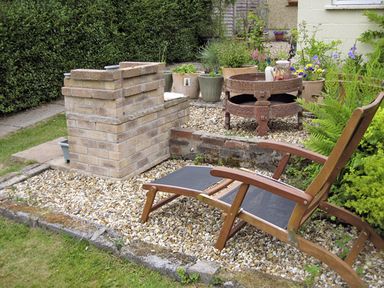 A garden area with a wooden lounge chair stone barbecue and potted plants at Lothlorien Cottage in Cwmgors near Ammanford