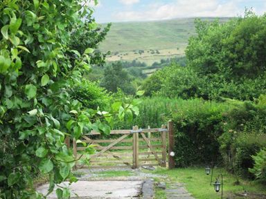 A garden path leading to a wooden gate surrounded by green bushes and trees with hills in the background at Lothlorien Cottage in Cwmgors near Ammanford
